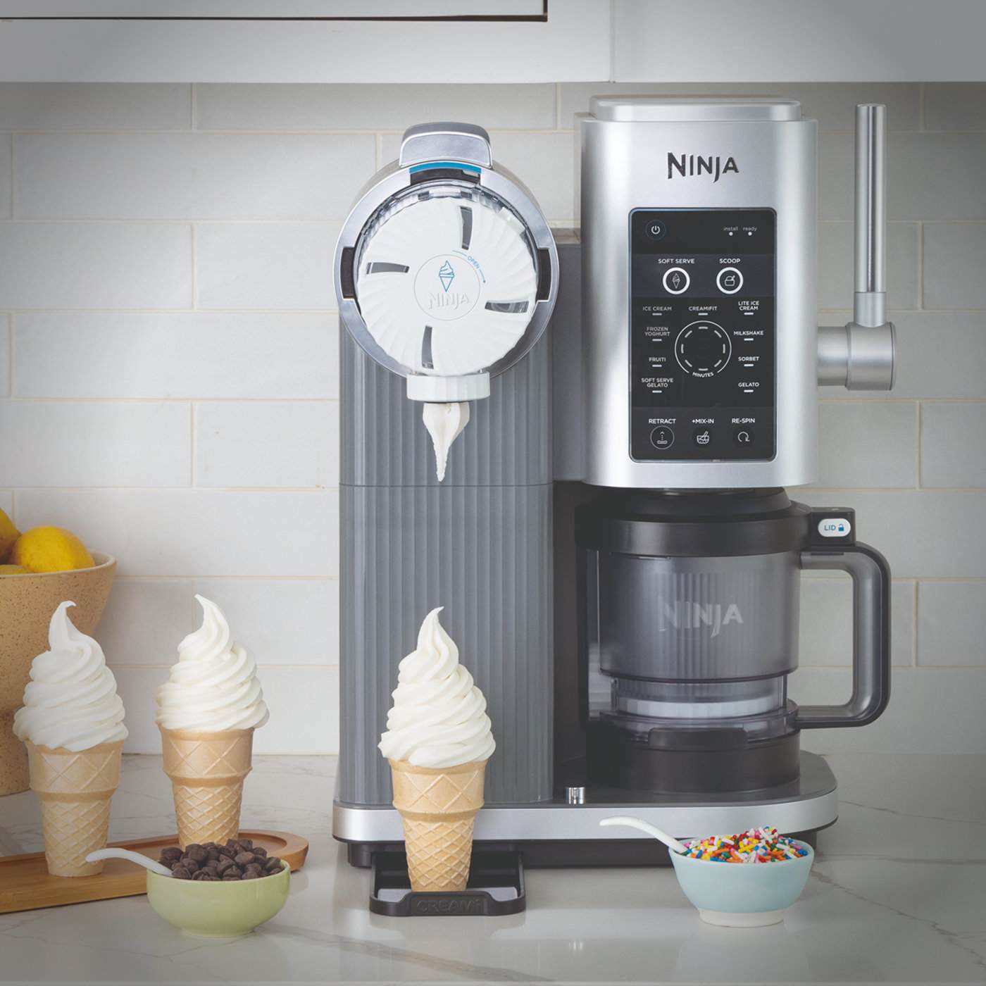 Ninja CREAMi dispensing soft serve into an ice cream cone, with cones and toppings arranged beside it.